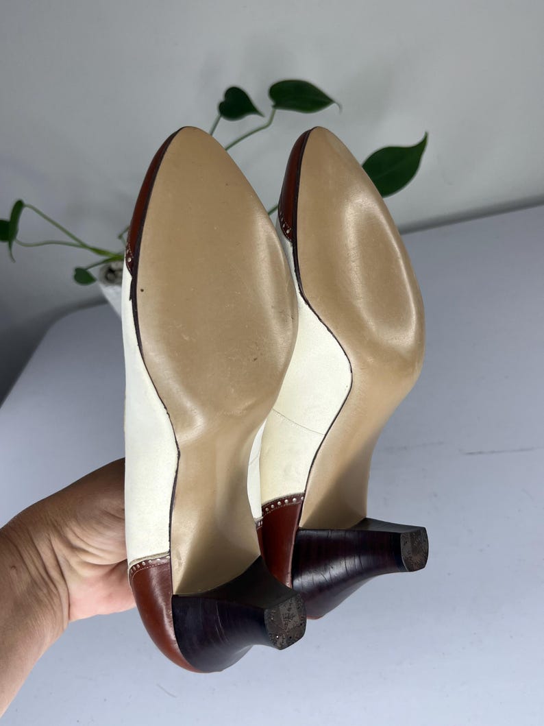 May include: A pair of vintage women's heels, viewed from the soles. The shoes feature a white upper with brown accents on the toe and heel. The heels are a dark brown, and the soles are a light tan color. The shoes have a classic, elegant design.