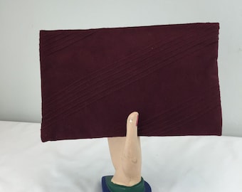 May include: A burgundy clutch with diagonal stitched lines. The clutch is rectangular and appears to be made of a soft fabric. It is displayed on a decorative hand-shaped stand with a blue, green, and beige base.