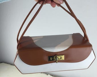 May include: A white and brown leather handbag with a gold clasp. The bag has a rectangular shape with rounded edges and two brown leather straps.