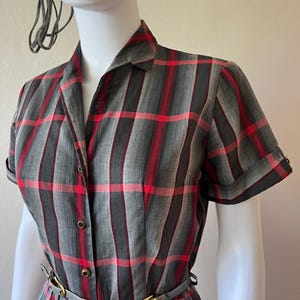 Peut inclure: Robe vintage &agrave; carreaux gris, rouges et noirs, avec manches courtes et col chemise. La robe a une taille ajust&eacute;e avec une fine ceinture et une jupe pliss&eacute;e. Pr&eacute;sent&eacute;e sur un mannequin.