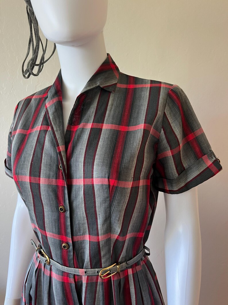 Peut inclure: Robe vintage &agrave; carreaux gris, rouges et noirs, avec manches courtes et col chemise. La robe a une taille ajust&eacute;e avec une fine ceinture et une jupe pliss&eacute;e. Pr&eacute;sent&eacute;e sur un mannequin.
