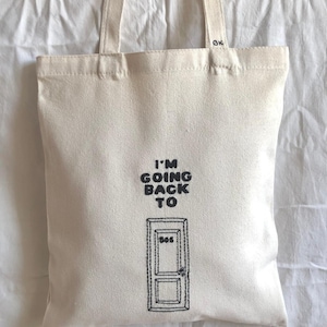 Può includere: Borsa tote in tela color crema con testo nero e un'illustrazione di una porta. Il testo recita "I'M GOING BACK TO" sopra un disegno di una porta con il numero "505". La borsa ha due manici e le iniziali "GK" sono visibili.