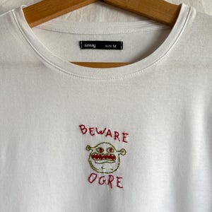 Op de afbeelding: Witte t-shirt met ronde hals met een geborduurd ontwerp van een ogre-gezicht en de woorden "BEWARE OGRE" in rode draad. Het shirt heeft een zwart label met de tekst "sinsay" en "SIZE M".