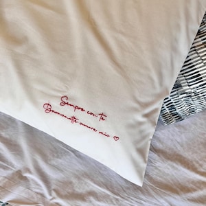 Può includere: Federa bianca con testo ricamato in rosso "Sempre con te, Buonanotte amore mio" e un simbolo a cuore. La federa è su un lenzuolo bianco, con un lenzuolo fantasia visibile sullo sfondo.