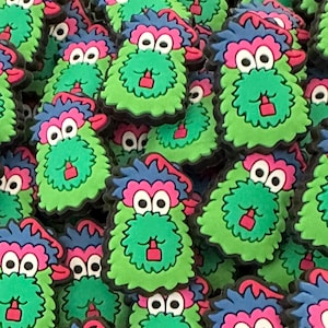 May include: A close-up of a pile of green, pink, and blue fuzzy monster charms. The monsters have two large eyes, a red mouth, and a fuzzy green body.