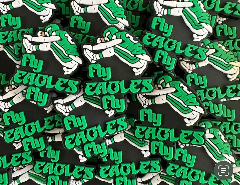 May include: A close-up of a pile of green and black rubber charms with the words "Fly Eagles Fly" printed on them. The charms are shaped like a football player in a green and white uniform.