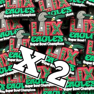 May include: A close-up of a pile of green and red Philadelphia Eagles Super Bowl Champions keychains. The keychains feature a stylized football helmet and the team's logo. The text "EAGLES Super Bowl Champions" is printed on the keychains. The image also includes the text "x2".