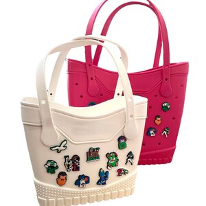 K&ouml;nnte beinhalten: Zwei Tragetaschen, eine wei&szlig;e und eine pinkfarbene, mit passenden Griffen. Die wei&szlig;e Tasche ist mit bunten Charms verziert. Die Taschen haben einen strukturierten Boden und bestehen aus einem gummiartigen Material. Die Taschen sind zum Tragen von Gegenst&auml;nden konzipiert.