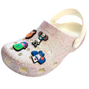 K&ouml;nnte beinhalten: Ein glitzernder rosa Clog-Schuh mit wei&szlig;em Innenraum und Riemen. Der Schuh ist mit bunten Cartoon-Charms verziert. Zu den Charms geh&ouml;ren ein gr&uuml;nes Monster, ein Footballspieler und ein blauer B&auml;r.