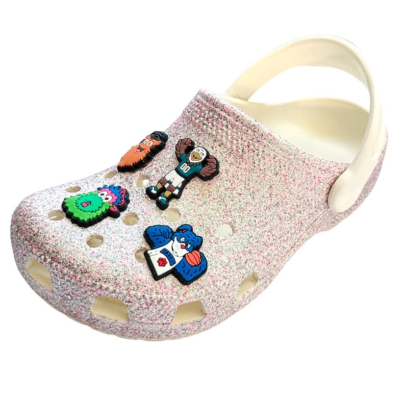 K&ouml;nnte beinhalten: Ein glitzernder rosa Clog-Schuh mit wei&szlig;em Innenraum und Riemen. Der Schuh ist mit bunten Cartoon-Charms verziert. Zu den Charms geh&ouml;ren ein gr&uuml;nes Monster, ein Footballspieler und ein blauer B&auml;r.