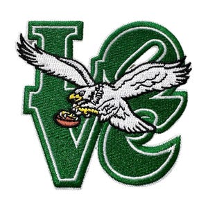May include: Embroidered patch featuring a white eagle with outstretched wings carrying a football. The eagle is positioned over the letters "VE" in green with white trim. The patch is designed for apparel or accessories.