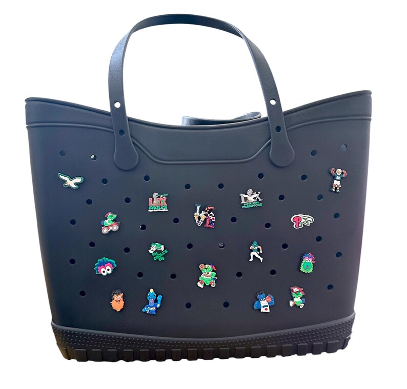 K&ouml;nnte beinhalten: Eine schwarze Tragetasche mit Griff und dekorativen Anh&auml;ngern. Die Tasche hat ein perforiertes Design und einen strukturierten Boden. Die Anh&auml;nger sind farbenfroh und zeigen verschiedene Sportmotive.