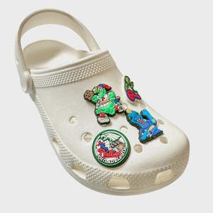 May include: White clog with a variety of colorful shoe charms. The charms include a green turtle, a blue and red guitar, a red cherry, a green leaf, and a round green and red Philadelphia Phillies logo.