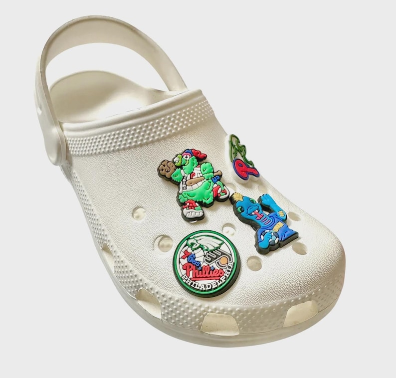 May include: White clog with a variety of colorful shoe charms. The charms include a green turtle, a blue and red guitar, a red cherry, a green leaf, and a round green and red Philadelphia Phillies logo.