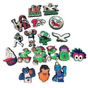 K&ouml;nnte beinhalten: Eine Sammlung bunter Schuhanh&auml;nger mit Logos und Maskottchen von Philadelphia-Sportteams. Zu den Anh&auml;ngern geh&ouml;ren die Eagles, Phillies und verschiedene Teammaskottchen. Die Anh&auml;nger sind aus Gummi und haben ein 3D-Design.