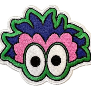 May include: Embroidered patch featuring a whimsical cartoon eye design. The patch has a white border, with green and blue accents around the pink eye shape. The eyes are white with black pupils. This patch is suitable for jackets, bags, or other fabric items.