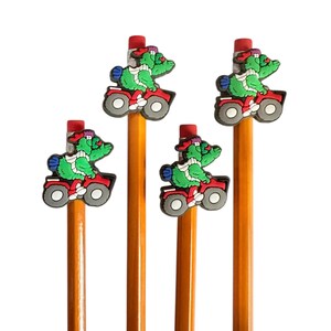 Puede incluir: Cuatro l&aacute;pices con gomas de borrar rojas y adornos de cocodrilo de dibujos animados montados en quads rojos. Los cocodrilos son verdes con bufandas blancas y sombreros rojos. Los l&aacute;pices son amarillos.