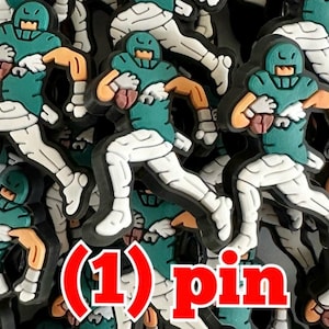 May include: A rubberized shoe charm featuring a football player in a teal jersey, white pants, and a helmet, holding a brown football while running. The figure has tan skin and white gloves. The text (1) pin is displayed in bold red letters.