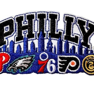 May include: An embroidered patch featuring the word PHILLY in black letters above a blue city skyline silhouette. Below the skyline are logos for Philadelphia sports teams including a red P, an eagle, a 76, a black and orange logo, and a gold circle.