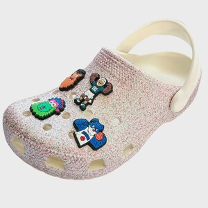 May include: A pair of white glitter clogs with five colorful shoe charms. The charms feature cartoon characters, including a green monster, a blue bear, a red-haired man, a blue bird, and a green and orange bird.