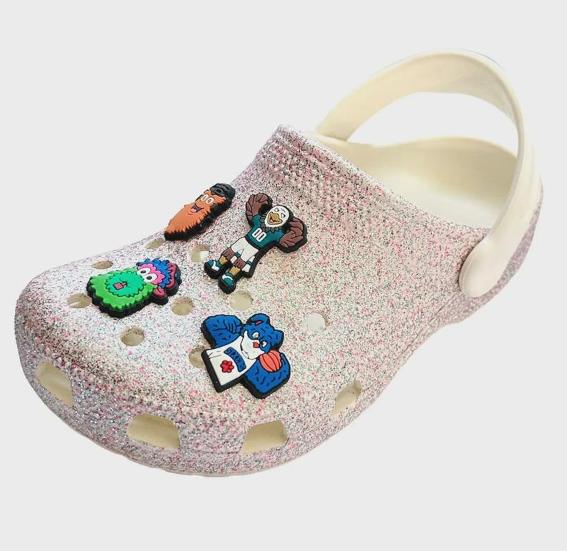 May include: A pair of white glitter clogs with five colorful shoe charms. The charms feature cartoon characters, including a green monster, a blue bear, a red-haired man, a blue bird, and a green and orange bird.
