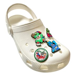 K&ouml;nnte beinhalten: Wei&szlig;e Clogs mit dekorativen Charms. Die Charms umfassen einen gr&uuml;nen und roten Baseballspieler, eine rote Kirsche, einen blauen Dinosaurier und ein Phillies-Logo. Die Clogs haben einen Riemen und Bel&uuml;ftungsl&ouml;cher.