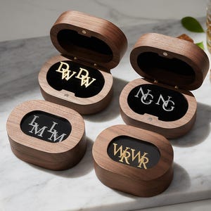 Op de afbeelding: Vier houten sieradendoosjes, elk met verschillende initialen in goud of zilver. De doosjes zijn open en onthullen de initialen op een zwarte fluwelen achtergrond. De doosjes staan op een marmeren oppervlak, met een glas amberkleurige vloeistof en ijsblokjes op de achtergrond.