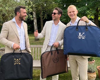 May include: Three garment bags in black, brown, and navy blue. The black bag features a gold monogram with arrows and a mustache. The brown bag has a black circular monogram with the letters GB. The navy bag displays the white text KERWIN.