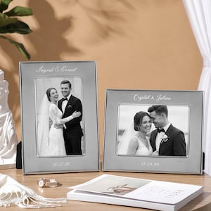 Pode incluir: Dois porta-retratos retangulares em tom prateado com fotografias de casamento em preto e branco. A moldura esquerda é vertical com o texto Ingrid & Ernest e 10.27.26. A moldura direita é horizontal com o texto Crystal & Julian e 11.25.26.