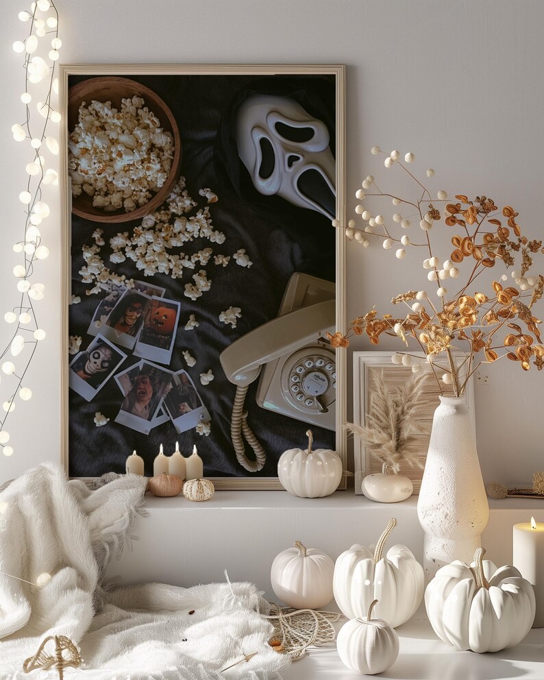 May include: A Halloween display with a framed image of a Scream mask, popcorn, and a vintage phone. White pumpkins, candles, and a vase with dried flowers create a spooky atmosphere. A string of fairy lights provides a soft illumination.