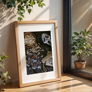 May include: Framed print with a horror movie theme. The image shows a Scream mask, popcorn, a vintage telephone, and small photos. The frame is light wood, and the background is a dark fabric.