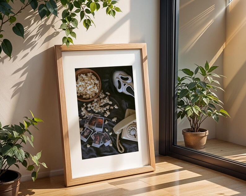 May include: Framed print with a horror movie theme. The image shows a Scream mask, popcorn, a vintage telephone, and small photos. The frame is light wood, and the background is a dark fabric.