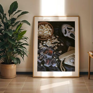 May include: A framed print with a horror movie theme. The image shows a bowl of popcorn, a Scream mask, a vintage telephone, and Polaroid photos. The scene is set on a black cloth, with a wooden frame and a light beige background.