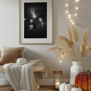 May include: A framed black and white print depicts two horror movie characters in a theater setting. The room is decorated with a bench, a cozy blanket, pumpkins, and a vase of pampas grass. A string of lights adds a warm glow.
