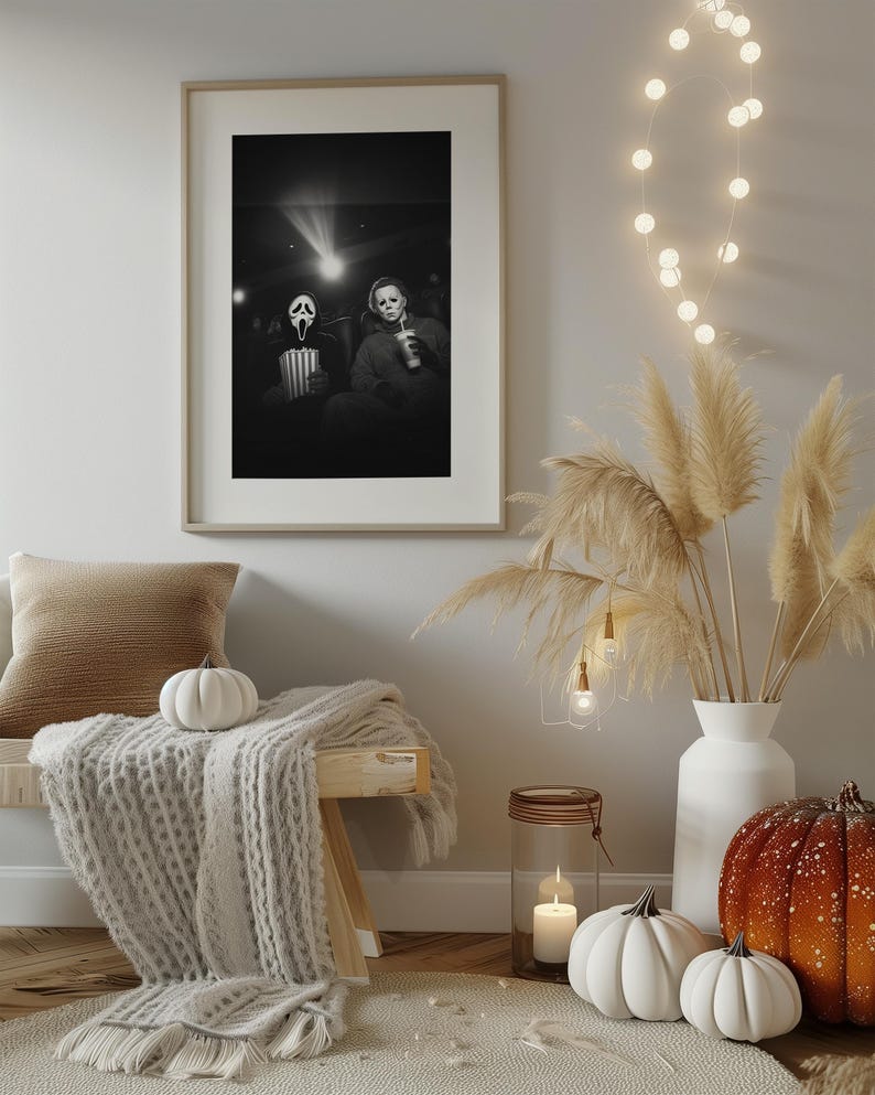 May include: A framed black and white print depicts two horror movie characters in a theater setting. The room is decorated with a bench, a cozy blanket, pumpkins, and a vase of pampas grass. A string of lights adds a warm glow.