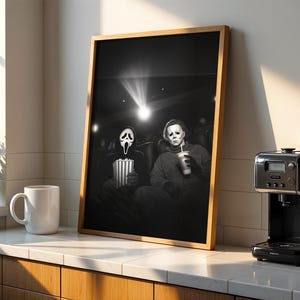 May include: Framed black and white print featuring two horror movie characters in a theater setting. One holds popcorn, the other a drink. The print is in a wooden frame, displayed on a countertop with a coffee maker and mug.