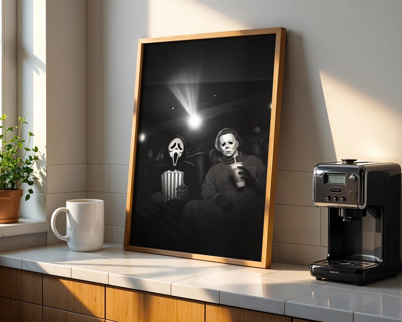 May include: Framed black and white print featuring two horror movie characters in a theater setting. One holds popcorn, the other a drink. The print is in a wooden frame, displayed on a countertop with a coffee maker and mug.