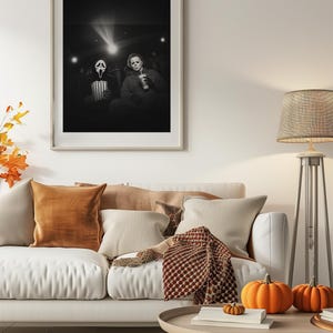 May include: A framed black and white photograph depicts two figures in a movie theater, one wearing a Ghostface mask and the other a Michael Myers mask. The room is decorated with a white sofa, orange pillows, pumpkins, and a floor lamp.
