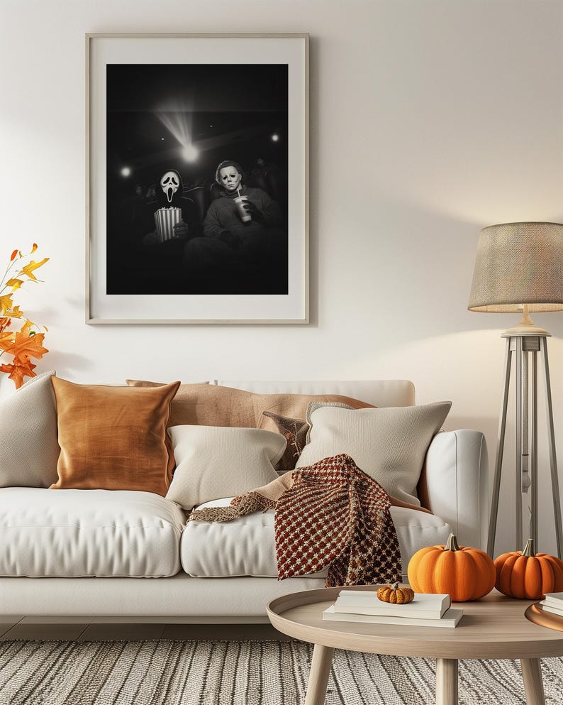 May include: A framed black and white photograph depicts two figures in a movie theater, one wearing a Ghostface mask and the other a Michael Myers mask. The room is decorated with a white sofa, orange pillows, pumpkins, and a floor lamp.