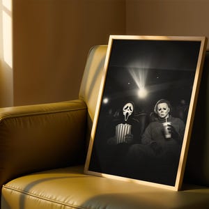 May include: A framed black and white print featuring two horror movie characters in a movie theater. One holds popcorn, the other a drink. The print has a light wood frame and is displayed on a green leather couch.