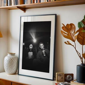 May include: Black and white framed print featuring two horror movie characters in a theater setting. One character wears a Scream mask and holds popcorn, the other wears a Michael Myers mask and holds a drink. The print is displayed on a white surface.