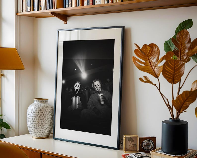 May include: Black and white framed print featuring two horror movie characters in a theater setting. One character wears a Scream mask and holds popcorn, the other wears a Michael Myers mask and holds a drink. The print is displayed on a white surface.