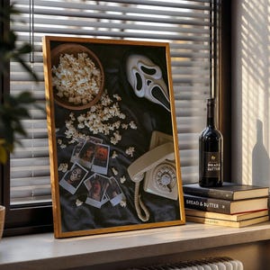 May include: A framed artwork with a horror movie theme, featuring a Scream mask, popcorn, and vintage photos. A bottle of wine and books are on a windowsill. The artwork is in a wooden frame.