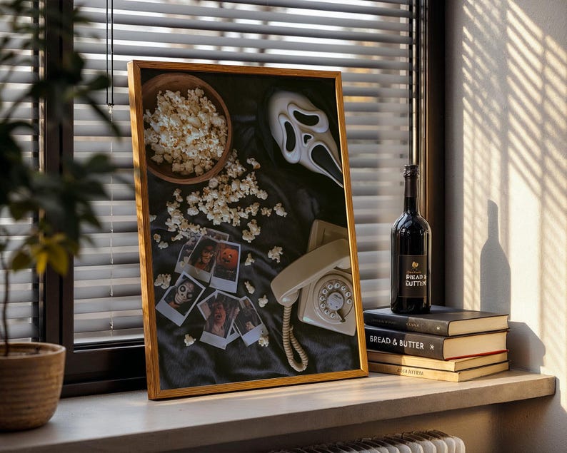 May include: A framed artwork with a horror movie theme, featuring a Scream mask, popcorn, and vintage photos. A bottle of wine and books are on a windowsill. The artwork is in a wooden frame.