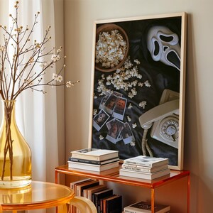 May include: A decorative arrangement with a framed artwork featuring a Scream mask, popcorn, and vintage photos. A yellow glass vase with branches and a retro telephone are also visible. The scene is set on a transparent orange table.