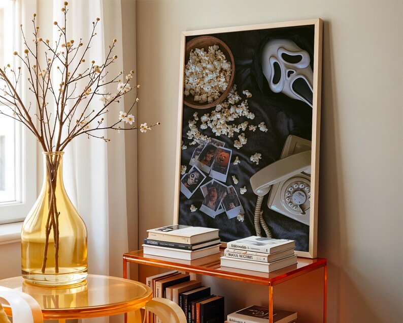 May include: A decorative arrangement with a framed artwork featuring a Scream mask, popcorn, and vintage photos. A yellow glass vase with branches and a retro telephone are also visible. The scene is set on a transparent orange table.