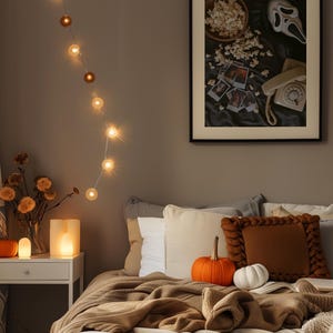 May include: A bedroom scene with a bed featuring a brown knitted blanket and pillows. An orange pumpkin and a white pumpkin are on the bed. A framed picture with popcorn and a Scream mask hangs on the wall. String lights provide a warm glow.