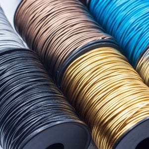 May include: Close-up of several spools of cord in various colors, including black, brown, gold, and blue. The cords are tightly wound on black spools, creating a textured, layered effect. The cords appear to be made of a shiny, smooth material.