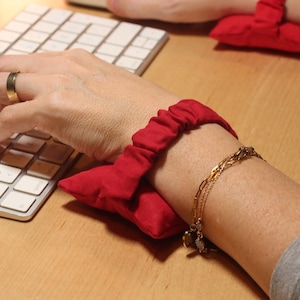 Op de afbeelding: Rode polssteun en steunkussen voor een computertoetsenbord. Het kussen is rechthoekig en gemaakt van een zachte, rode stof. De polssteun is ontworpen om ondersteuning en comfort te bieden tijdens het typen. Een gouden armband is ook zichtbaar.