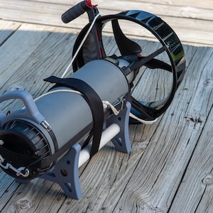 May include: A gray and black underwater propulsion device with a handle, a propeller guard, and a cylindrical body. The device is supported by a light blue frame and secured with black straps. It rests on a weathered wooden surface.